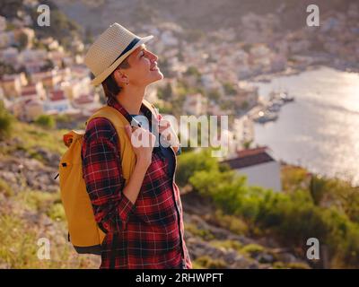 Bella donna asiatica felice con zaino che si gode le vacanze alle isole Symi al tramonto. Vista del porto di Symi o Simi, è una piccola isola del Dodecaneso, Grecia, atmosfera tranquilla e architettura favolosa. Foto Stock
