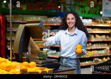 Ritratto di una donna soddisfatta del supermercato, donna ispanica che pesa frutta in negozio, sorride e guarda la fotocamera utilizzando bilance self-service. Foto Stock