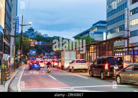 Il traffico attende ad un incrocio di sera sulla trafficata Densha Dori vicino alla fermata del tram Torichosuji, con il castello di Kumamoto che torreggia. Foto Stock