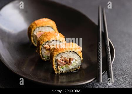 Fila concentrata di gustosi panini fritti di sushi con riso cotto e salmone con fette di avocado su un piatto nero di ceramica sul tavolo Foto Stock