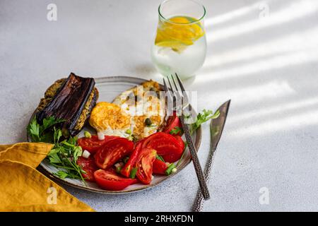 Dall'alto del piatto con un delizioso pranzo salutare che include uova fritte con fette di pomodoro, prezzemolo, cipolla primaverile, capperi serviti con un bicchiere di pu freddo Foto Stock
