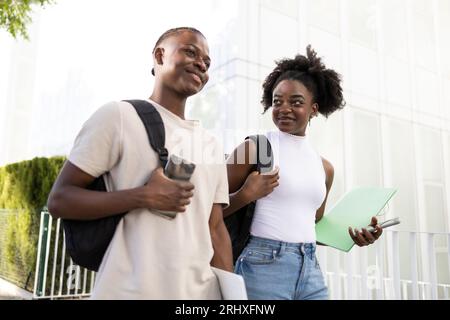 Dal basso, allegra coppia di studenti afro-americani adolescenti in abiti casual con zaini sorridenti e guardando lontano mentre cammini su strada lastricata Foto Stock