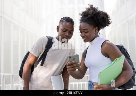 Coppia adolescente afroamericana concentrata in abiti informali con zaini che naviga sul cellulare mentre si trova in strada insieme di giorno Foto Stock