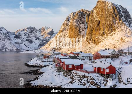 Spettacolare vista aerea di piccole case illuminate dalla luce del sole situate su una spiaggia innevata contro le montagne rocciose delle Isole Lofoten in Norvegia Foto Stock