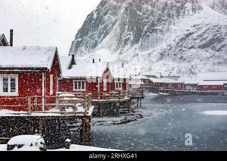 Maestoso paesaggio di piccoli insediamenti di case rosse situate sulla riva del fiume con il tempo innevato contro le alte montagne in inverno sulle isole Lofoten a NOR Foto Stock