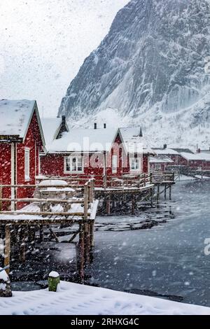 Maestoso paesaggio di piccoli insediamenti di case rosse situate sulla riva del fiume con il tempo innevato contro le alte montagne in inverno sulle isole Lofoten a NOR Foto Stock