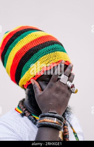 Anonimo afroamericano con un cappello colorato che si staglia e guarda lontano mentre copre il viso con la mano su sfondo bianco Foto Stock
