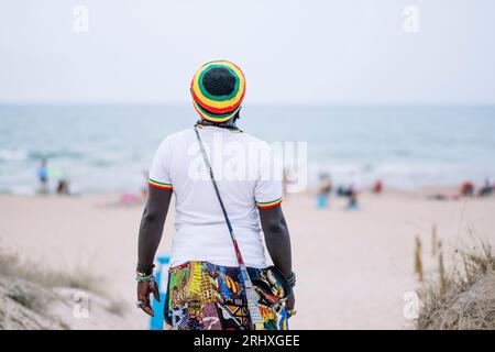 Vista posteriore di un anonimo uomo afroamericano con abiti colorati e cappello in piedi contro una spiaggia sfocata Foto Stock
