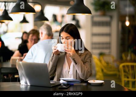 Giovane e tranquilla freelance seduta a un tavolo con un computer portatile e bevendo un caffè mentre lavora a un progetto remoto in un bar con sfondo sfocato delle persone Foto Stock