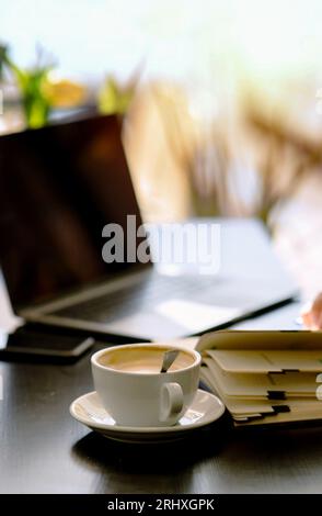Messa a fuoco selettiva di caffè cremoso in tazza con cucchiaio e notebook sul tavolo con sfondo sfocato di laptop e telefono aperti al bar alla luce del giorno Foto Stock