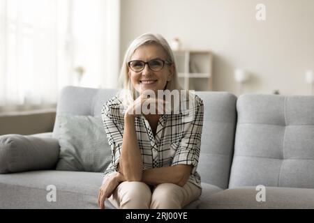 Donna matura bionda felice con eleganti occhiali da vista seduti sul divano Foto Stock