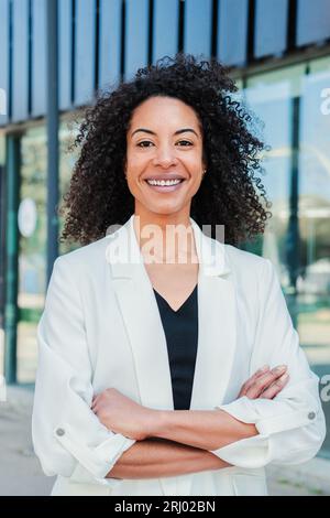 Ritratto verticale di una donna d'affari con braccia ispaniche incrociate con i capelli ricci che sorridono guardando la fotocamera e che si trova in piedi sullo spazio di lavoro indossando un abito. Dipendente aziendale piegato con un atteggiamento di successo. Foto di alta qualità Foto Stock