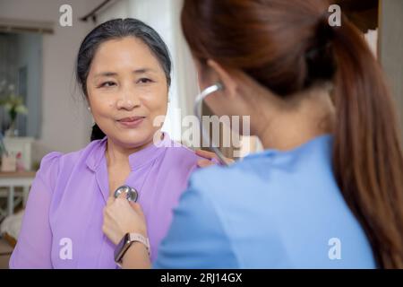 Giovane infermiera asiatica o infermiera seduto sul divano tenendo stetoscopio ascoltare il battito cardiaco con la donna anziana nel soggiorno di casa, controllare il medico cardiologo Foto Stock