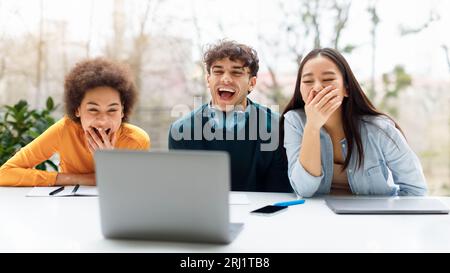 Allegri amici di studenti diversi che guardano contenuti divertenti insieme sul laptop, seduti in casa e ridendo, panorama Foto Stock