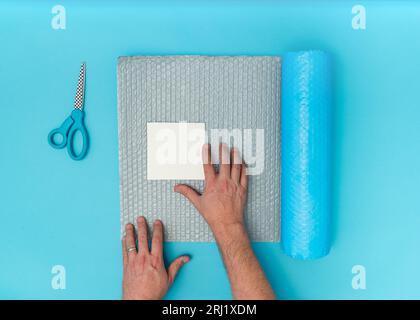 Vista dall'alto delle mani maschili adulti caucasiche che confezionano una carta quadrata bianca con involucro a bolle d'aria e forbici. Sfondo blu con spazio di copia. Foto Stock