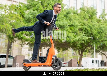 Un uomo d'affari eccitato e divertente in completo a cavallo di uno scooter durante una riunione d'affari. Uomo d'affari che guida uno scooter elettrico su strada in città mentre c'è traffico Foto Stock