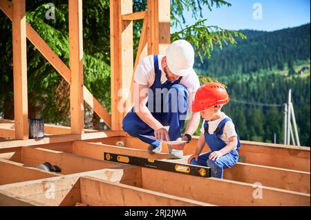 Padre con figlio piccolo che costruisce una casa con struttura in legno. Lavoratore maschile che mostra al figlio il piano di costruzione, indossando caschi e tute blu nelle giornate di sole. Carpenteria e concetto di famiglia. Foto Stock