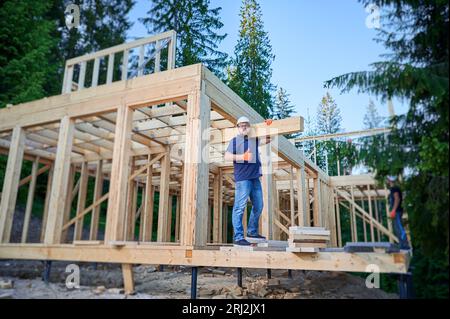 Carpenter costruisce una casa con struttura in legno vicino alla foresta. L'uomo barbuto tiene una grande tavola sulla spalla, mostrando i pollici in alto, indossando abiti da lavoro e casco. Concetto di moderna costruzione ecologica. Foto Stock