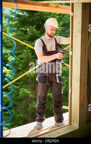 Falegname costruzione casa in legno incorniciato. Uomo bearded lavoro rivestimento facciata di casa, allacciatura con cacciavite, indossare tute da lavoro e casco. Concetto di moderna costruzione ecologica. Foto Stock