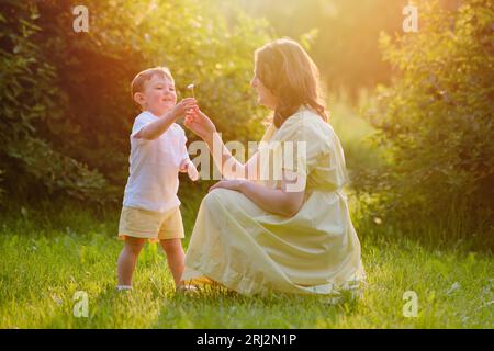 Una madre donna dà un fiore a un bambino in natura. Un bambino felice con la mamma in una passeggiata nel parco estivo. Bambino di circa due anni (un anno e undici mesi Foto Stock