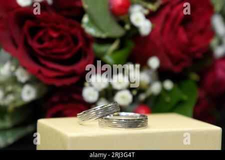 Un primo piano di due fedi nuziali poggiati su una piccola scatola bianca, con un bouquet di rose rosse sullo sfondo Foto Stock