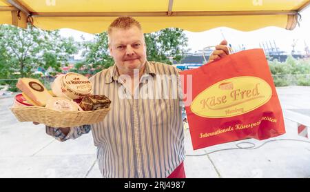 Amburgo, Germania. 23 luglio 2023. Axel Pabst vende formaggio al mercato del pesce di Amburgo. Da più di 25 anni Axel Pabst lavora come market crier per l'azienda Fred4Food, che è diventata nota nel mercato del pesce attraverso il commercio di frutta Bananen-Fred. Nel frattempo, però, non c'è più solo frutta nell'assortimento, ma anche, tra le altre cose, il proprio stand di formaggi. Meglio conosciuto come: Cheese-Fred. (A dpa: "Gridare per Käse-Fred - un venditore di pesce in missione") credito: Markus Scholz/dpa/Alamy Live News Foto Stock