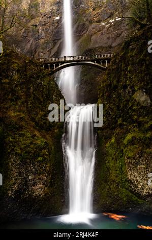 Una maestosa cascata che scende giù da una lussureggiante scogliera rocciosa ricoperta di muschio, con un ponte che si arrocca sull'acqua turbolenta Foto Stock