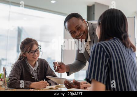 Un felice insegnante o tutor di inglese afroamericano sta ridendo e divertendosi ad insegnare agli studenti asiatici in classe in una scuola di lingue. Istruzione e mu Foto Stock