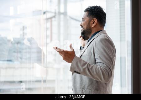 Un uomo d'affari indiano-asiatico professionista e di successo o un amministratore delegato maschile in una tuta formale sta parlando al telefono mentre si trova accanto alla finestra in un Foto Stock