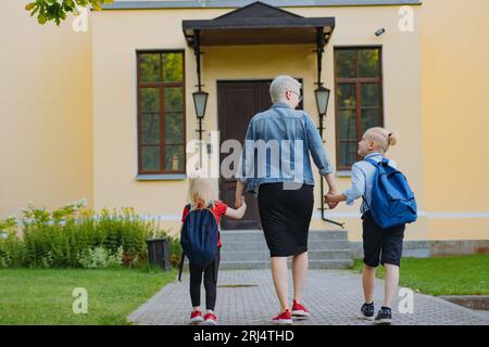 la madre accompagna i bambini a scuola tenendosi per mano. Bionda caucasica con i capelli legati in coda di cavallo e bambina che va a scuola con la mamma. Imag Foto Stock