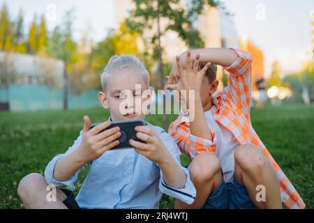 simpatici e divertenti bambini che scattano selfie con lo smartphone che crea una cornice di dita. I bambini nel parco seduti sull'erba si divertono insieme. Immagine con f. Selettiva Foto Stock