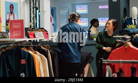 Persone diverse che si affrettano a scegliere i vestiti durante il grande evento di vendita del Black friday, correndo in un negozio di abbigliamento per ottenere le migliori offerte. Una folla aggressiva di acquirenti che acquistano capi di abbigliamento. Scatto con treppiede. Foto Stock