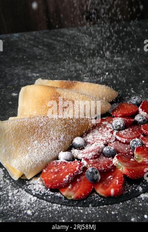 foto frittelle sottili e tante bacche fresche cosparse sopra di zucchero a velo su uno sfondo scuro di grandi dimensioni Foto Stock