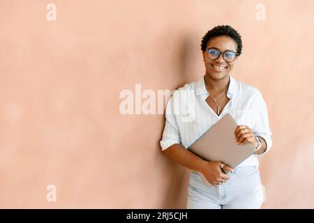 Brillante giovane donna d'affari africana in occhiali casual sorridente con un dente di fessura. Allegra e felice studentessa nera che porta un notebook, guarda la fotocamera e ride, pronta per una giornata produttiva Foto Stock