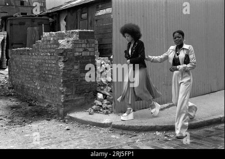 Moda anni '1970, due donne britanniche nere. Due amiche, giovani donne nere alla moda che indossano scarpe con suola con plateau e una con un paio di pantaloni. Brick Lane, Whitechapel, Londra circa giugno 1974 HOMER SYKES 1 Foto Stock