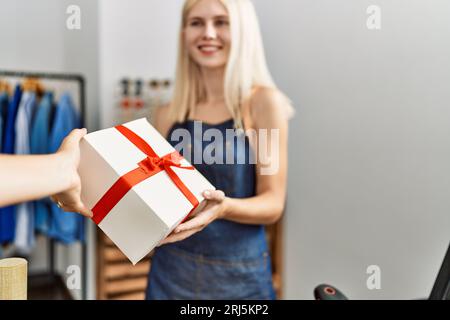 Giovane assistente del negozio bionda sorridente sicura di offrire un regalo al cliente in un negozio di abbigliamento Foto Stock