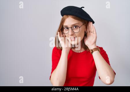 Giovane donna rossa in piedi con occhiali e berretto che cerca di sentire entrambe le mani sul gesto dell'orecchio, curiosa di pettegolezzi. problemi all'udito, sordo Foto Stock