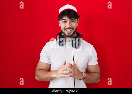 Uomo ispanico con barba che indossa cappello da giocatore e cuffie mani unite e dita incrociate sorridendo rilassato e allegro. successo e ottimismo Foto Stock