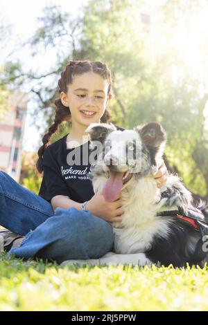 Ritratto verticale di una bambina dai capelli rossi con trecce e lentiggini sorridenti, seduta nel parco con il suo cane bianco e nero Foto Stock