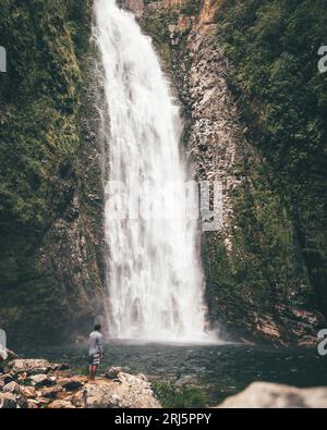 Un giovane uomo si trova vicino a una maestosa cascata che scende lungo un lussureggiante paesaggio tropicale della giungla Foto Stock