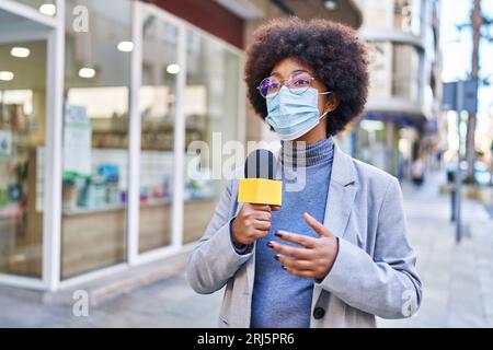 Donna afroamericana reporter indossare maschera medica utilizzando il microfono che lavora per strada Foto Stock