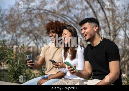 Tre giovani multirazziali che inviano SMS con smartphone seduti in un parco. Gruppo eterogeneo che sorride e usa i telefoni all'aperto seduti fuori. Foto Stock