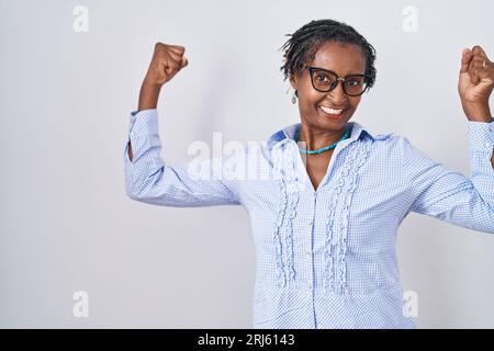 Donna africana con dreadlocks su sfondo bianco che indossa occhiali che mostrano i muscoli delle braccia sorridendo orgogliosa. concetto di fitness. Foto Stock