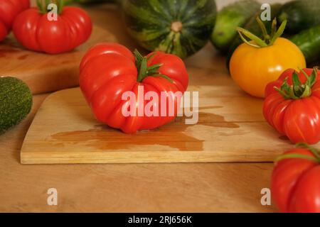 Un pomodoro grande maturo fresco su un tagliere di legno. Molte verdure crude di fondo. Pomodori, cetrioli. Fornello a casa che fa un pasto vegetariano sano. Foto Stock