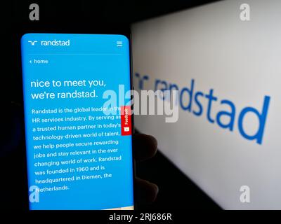 Persona in possesso di cellulare con pagina web dell'azienda olandese di risorse umane Randstad N.V. sullo schermo con logo. Concentrarsi sul centro del display del telefono. Foto Stock