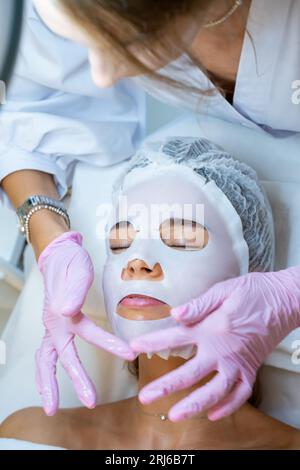 Cosmetologo che indossa guanti di gomma applicazione maschera foglio sul viso della donna. Foto Stock