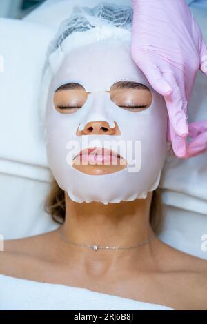 Cosmetologo che indossa guanti di gomma applicazione maschera foglio sul viso della donna. Foto Stock