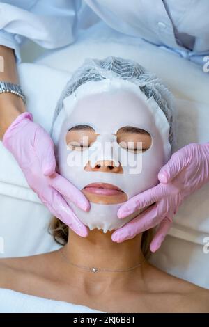 Cosmetologo che indossa guanti di gomma applicazione maschera foglio sul viso della donna. Foto Stock