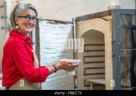 Artigiana che colloca tazze di argilla nel forno per cuocere la ceramica mentre lavora in officina. Foto Stock