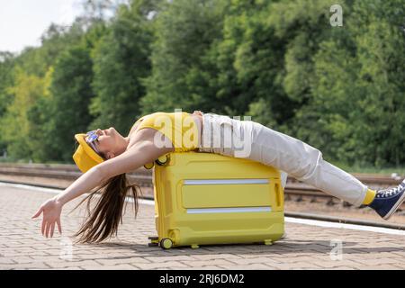 Bacio del vento: Un viaggiatore con un cappotto giallo è pronto a rilassarsi all'aria aperta. Una donna è sdraiata sulla sua valigia, in attesa di essere trasportata o l'ha appena fatto Foto Stock